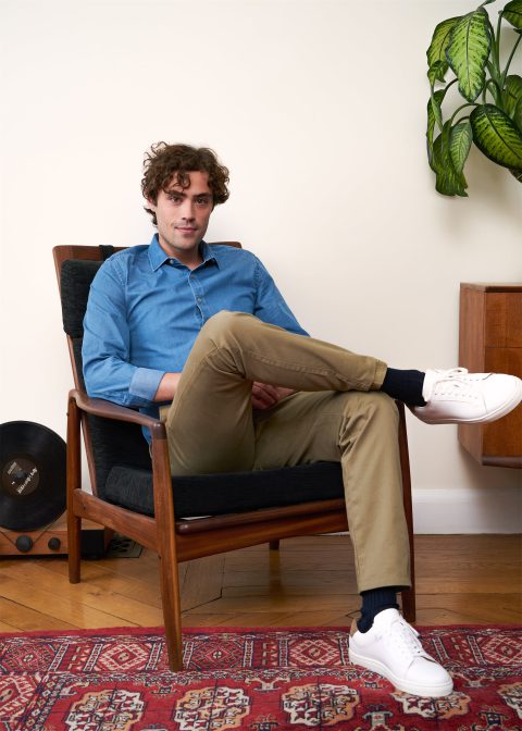Image de campagne pour Bexley montrant un homme assis dans un fauteuil en bois au style vintage, vêtu d'une chemise bleue et d'un pantalon beige, posant dans un intérieur cosy avec un tapis persan rouge et une plante verte en arrière-plan, il est fier et détendu