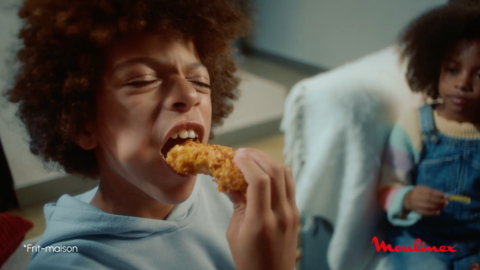 Extrait de la vidéo Moulinex montrant un enfant croqué dans un bout de poulet frit doré. On devine sa soeur en arrière plan. La scène est gourmande et détendue.